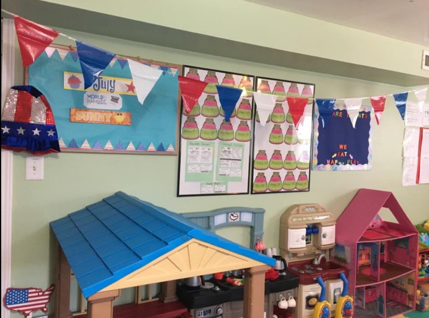 A play kitchen and dollhouse corner beneath a July-themed bulletin board