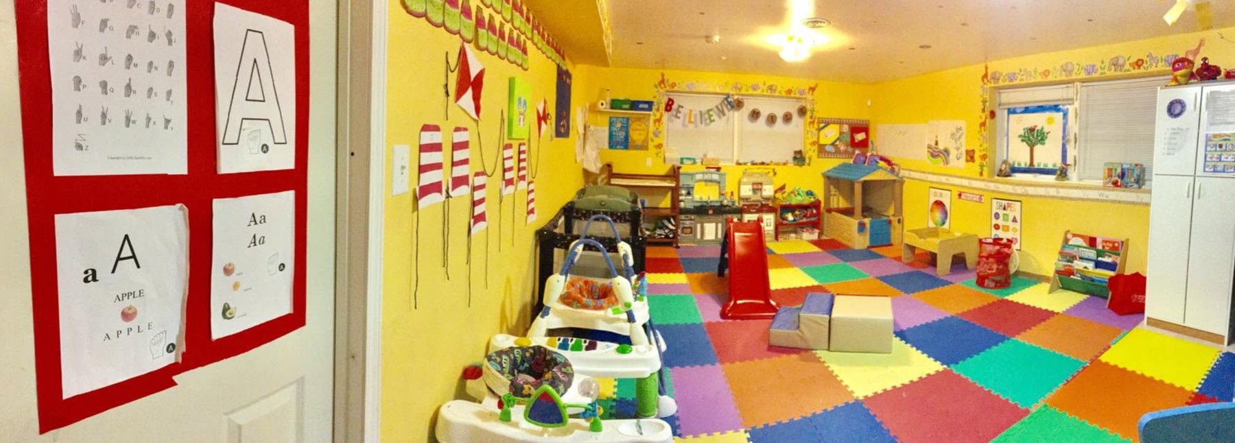A bright infant and toddler room with a sign-language alphabet poster on the door and a colorful foam play mat